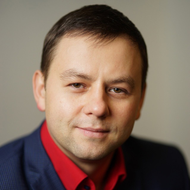Close-up portrait of a man in a red shirt and dark blazer, looking at the camera with a gentle smile (professional headshot).