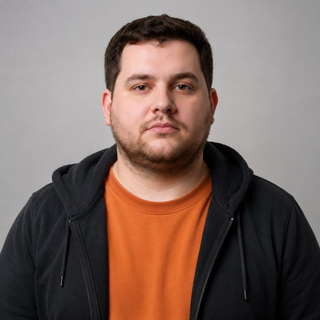Studio portrait of a man with short dark hair and stubble, wearing an orange shirt under a black hoodie.